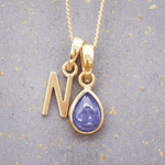 Gold necklace with 'N' initial pendant and teardrop-shaped purple december birthstone tanzanite gemstone on a textured grey background.