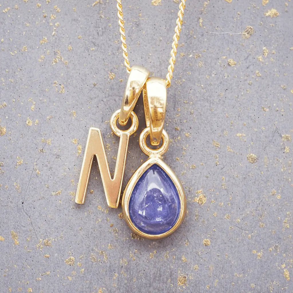 Gold December birthstone necklace with 'N' initial pendant and a natural tanzanite gemstone pendant on a grey background.