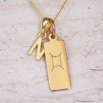 Gold initial N and Gemini zodiac bar necklace, close-up of the letter charm and engraved pendant on whitewashed wood