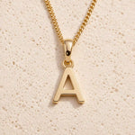 Personalised gold initial letter pendant necklace with polished uppercase A pendant on a fine cable chain in 9k gold plated sterling silver against a textured beige stone background