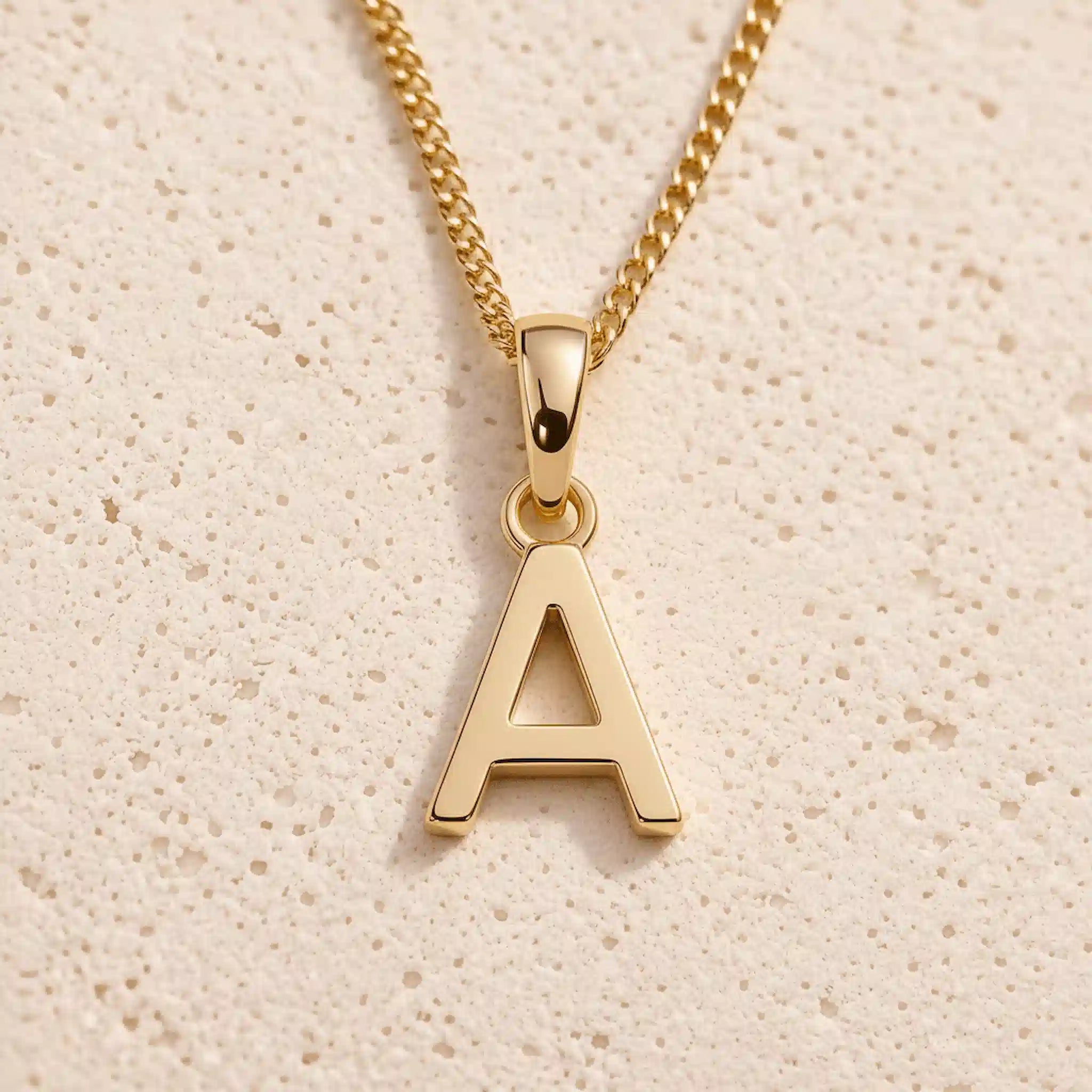 Personalised gold initial letter pendant necklace with polished uppercase A pendant on a fine cable chain in 9k gold plated sterling silver against a textured beige stone background