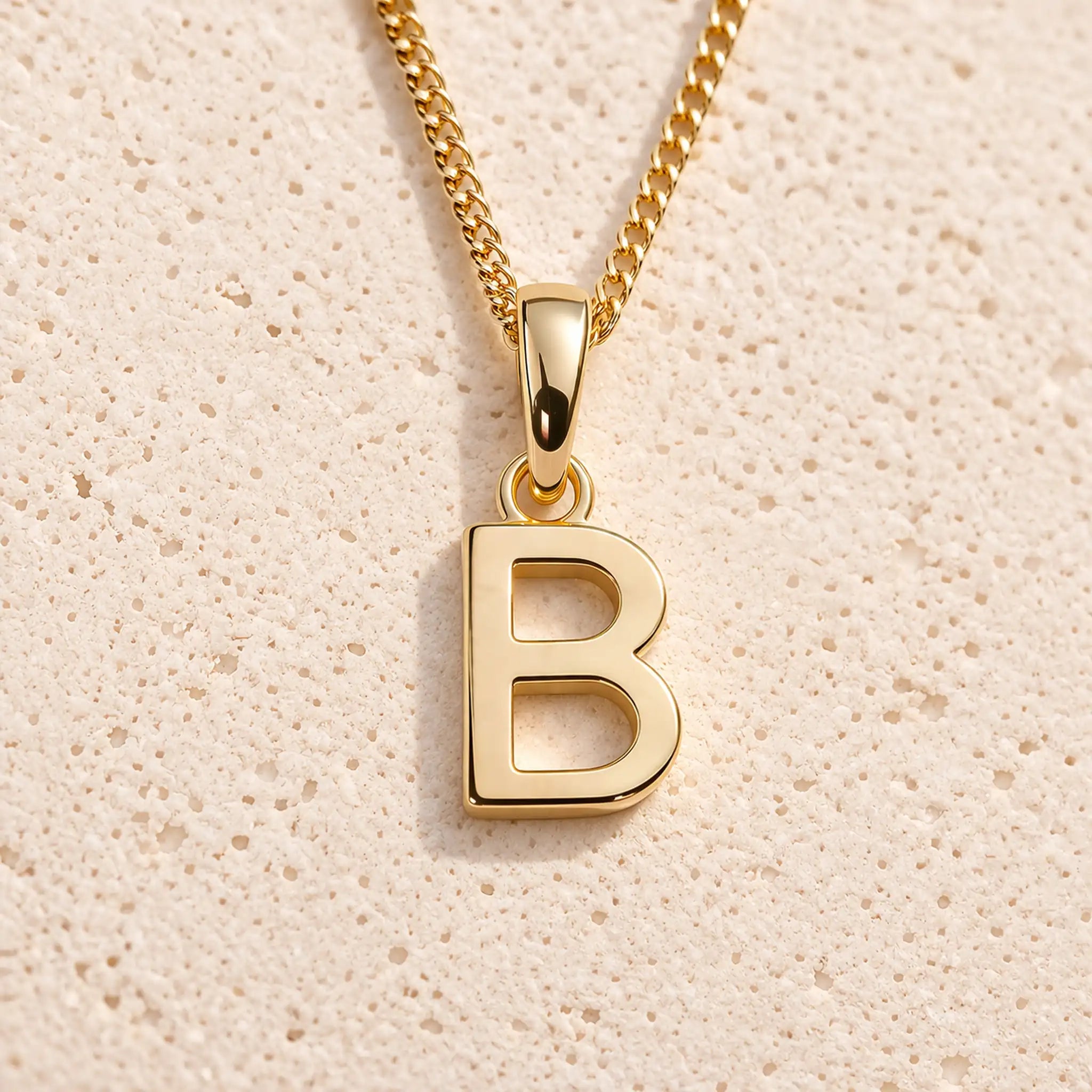 Personalised gold initial letter pendant necklace with polished uppercase B pendant on a fine cable chain in 9k gold plated sterling silver against a textured beige stone background