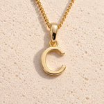 Personalised gold initial letter pendant necklace with polished uppercase C pendant on a fine cable chain in 9k gold plated sterling silver against a textured beige stone background