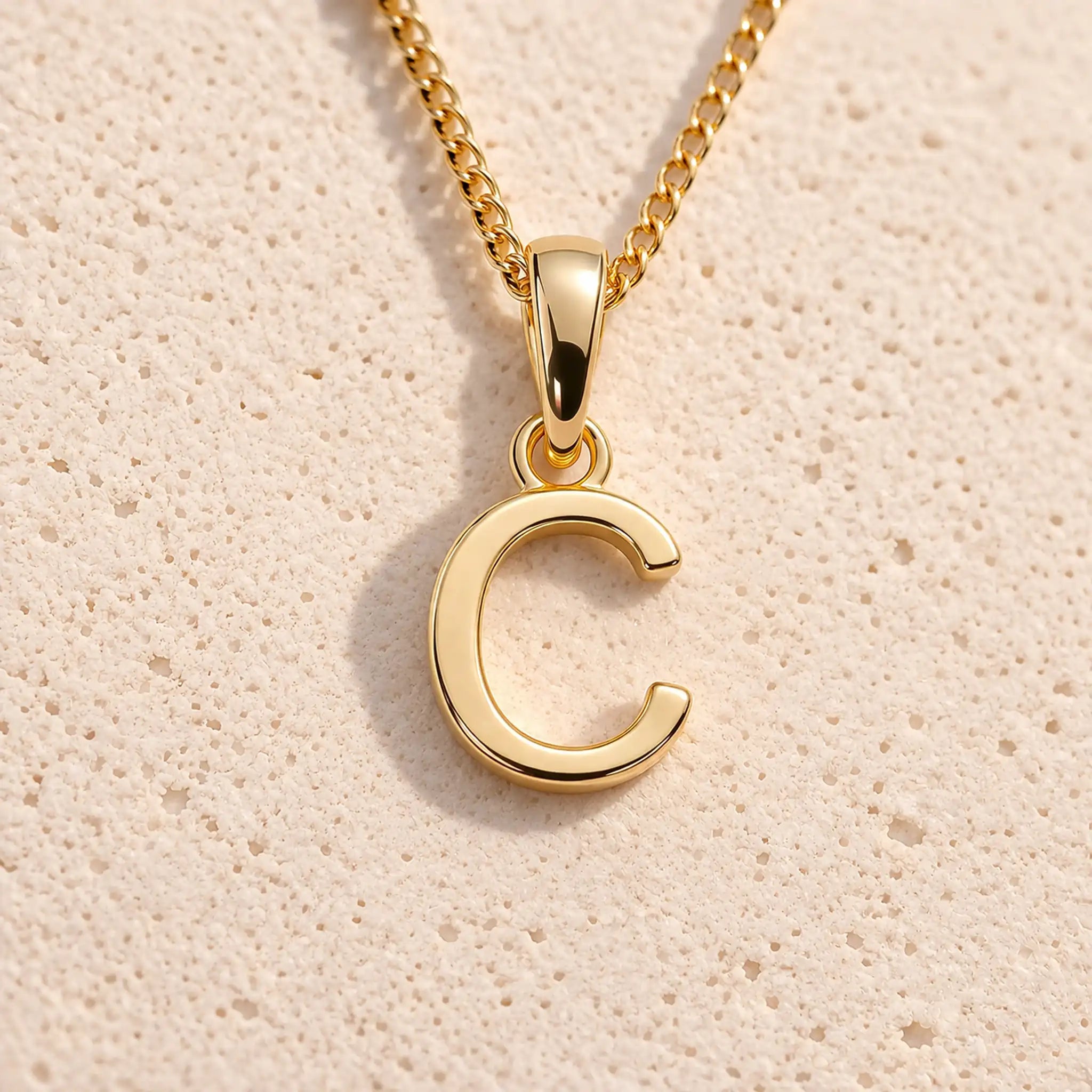 Personalised gold initial letter pendant necklace with polished uppercase C pendant on a fine cable chain in 9k gold plated sterling silver against a textured beige stone background