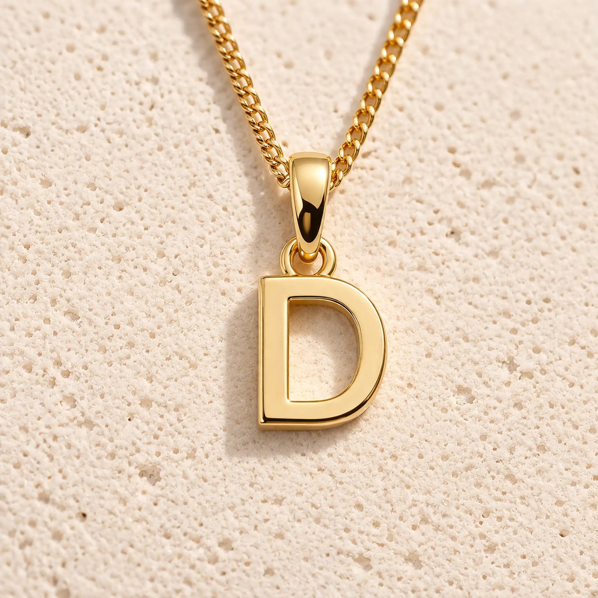 Personalised gold initial letter pendant necklace with polished uppercase D pendant on a fine cable chain in 9k gold plated sterling silver against a textured beige stone background
