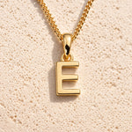 Personalised gold initial letter pendant necklace with polished uppercase E pendant on a fine cable chain in 9k gold plated sterling silver against a textured beige stone background
