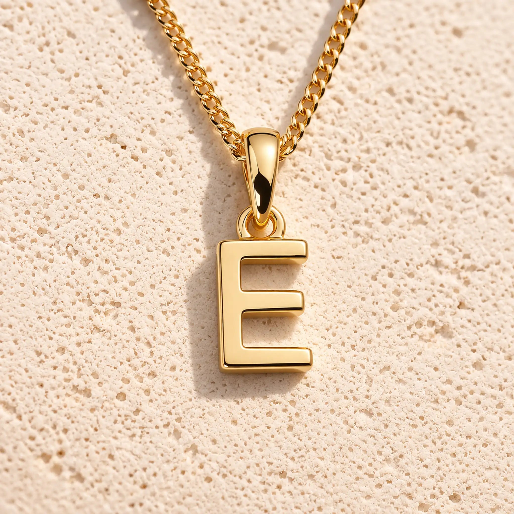 Personalised gold initial letter pendant necklace with polished uppercase E pendant on a fine cable chain in 9k gold plated sterling silver against a textured beige stone background
