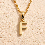 Personalised gold initial letter pendant necklace with polished uppercase F pendant on a fine cable chain in 9k gold plated sterling silver against a textured beige stone background