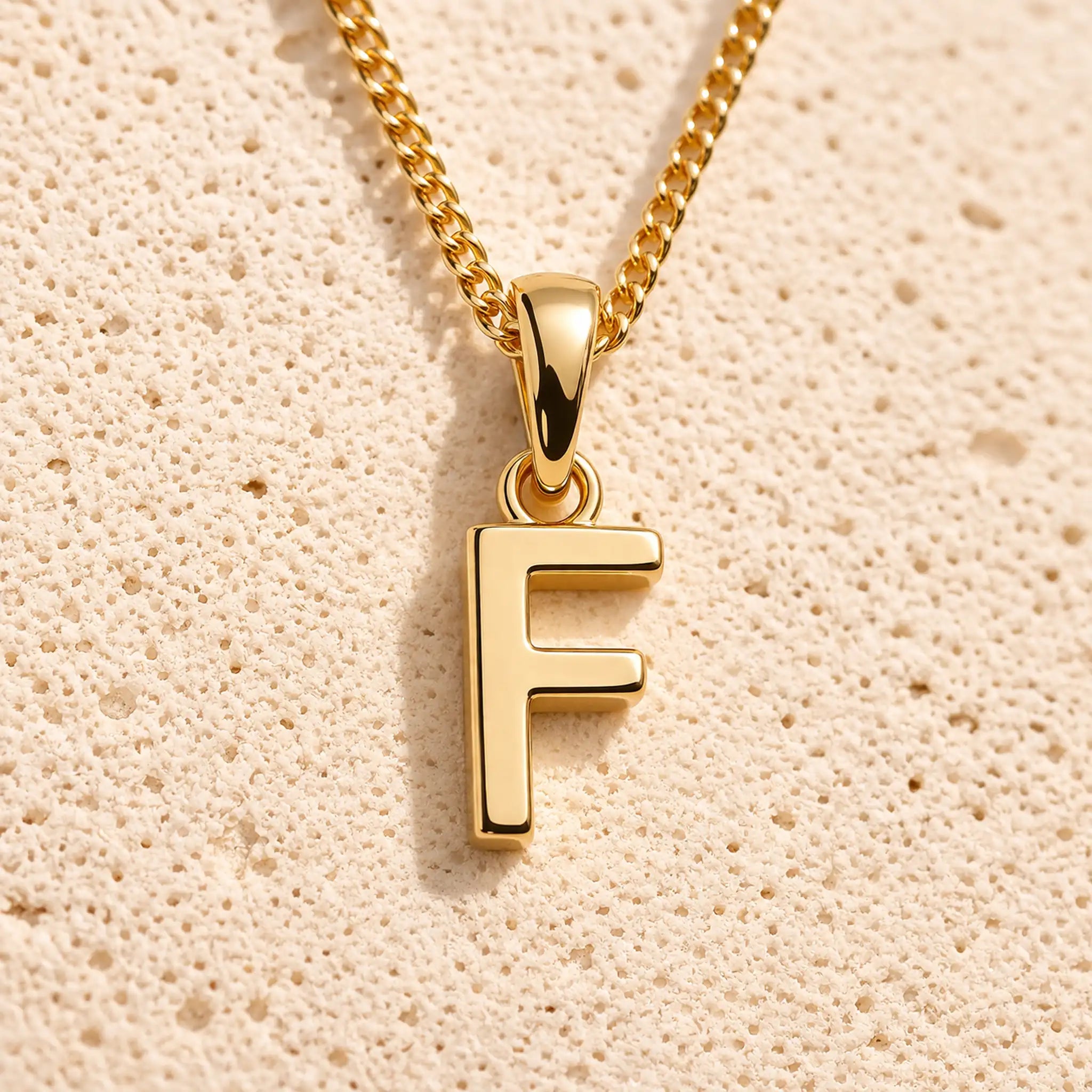 Personalised gold initial letter pendant necklace with polished uppercase F pendant on a fine cable chain in 9k gold plated sterling silver against a textured beige stone background