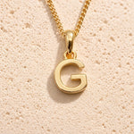 Personalised gold initial letter pendant necklace with polished uppercase G pendant on a fine cable chain in 9k gold plated sterling silver against a textured beige stone background