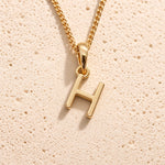 Personalised gold initial letter pendant necklace with polished uppercase H pendant on a fine cable chain in 9k gold plated sterling silver against a textured beige stone background