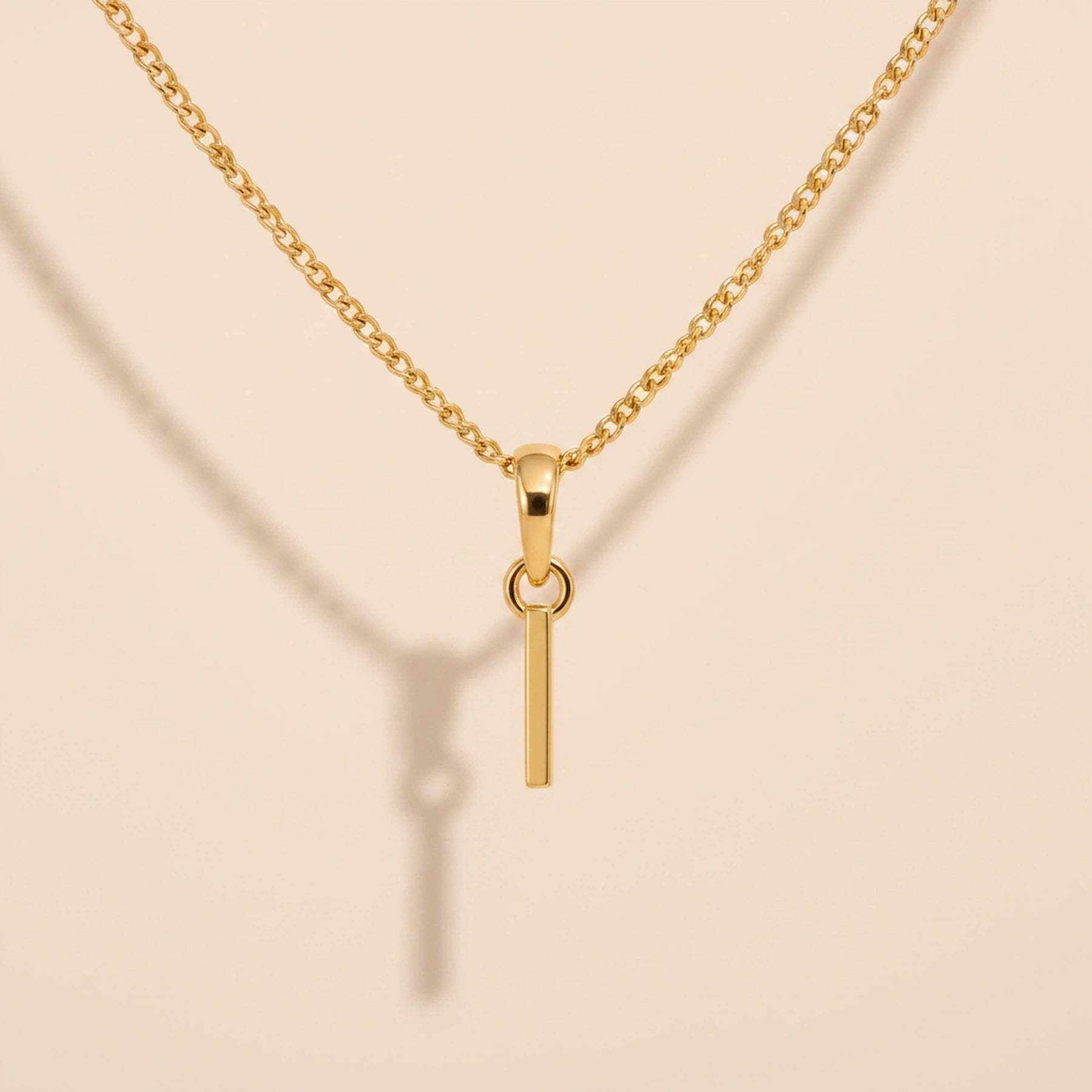 Personalised gold initial letter pendant necklace with polished uppercase I pendant on a fine cable chain in 9k gold plated sterling silver against a textured beige stone background