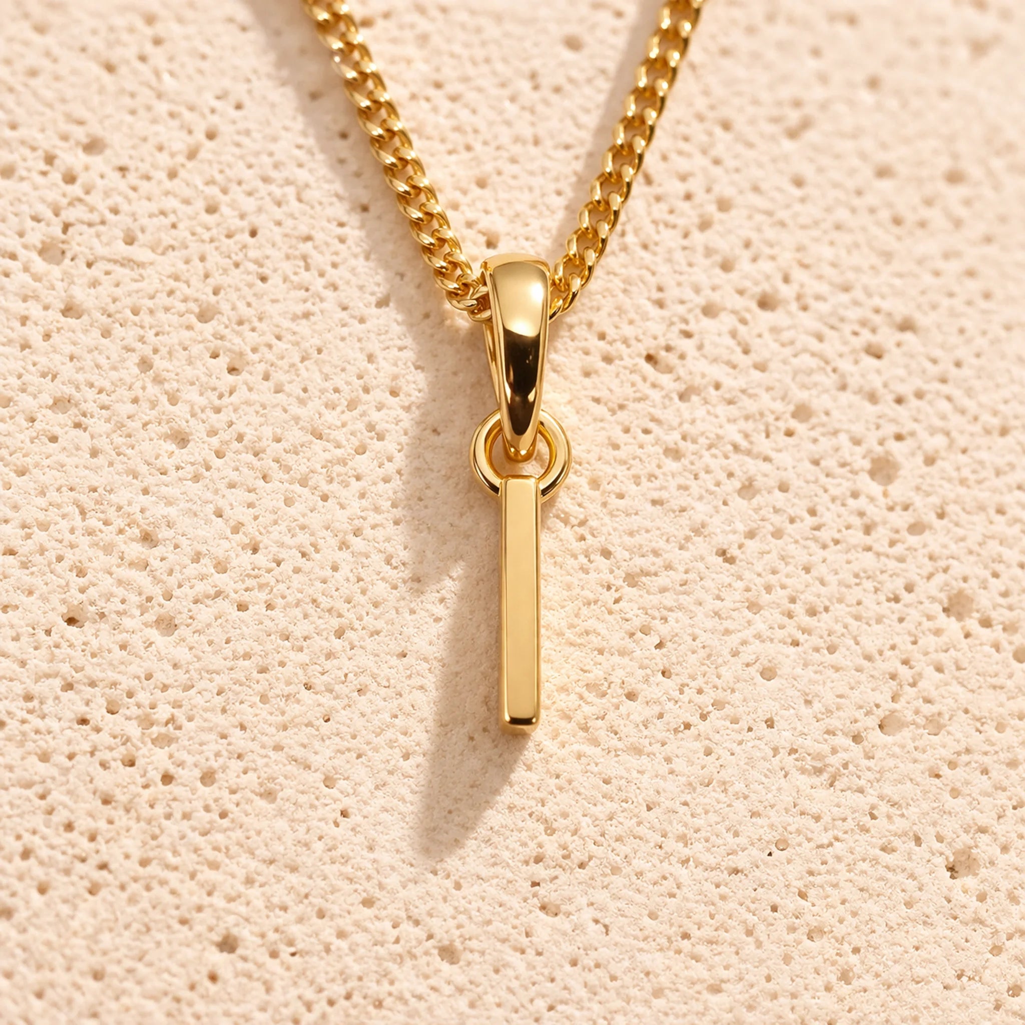 Personalised gold initial letter pendant necklace with polished uppercase I pendant on a fine cable chain in 9k gold plated sterling silver against a textured beige stone background
