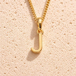 Personalised gold initial letter pendant necklace with polished uppercase J pendant on a fine cable chain in 9k gold plated sterling silver against a textured beige stone background