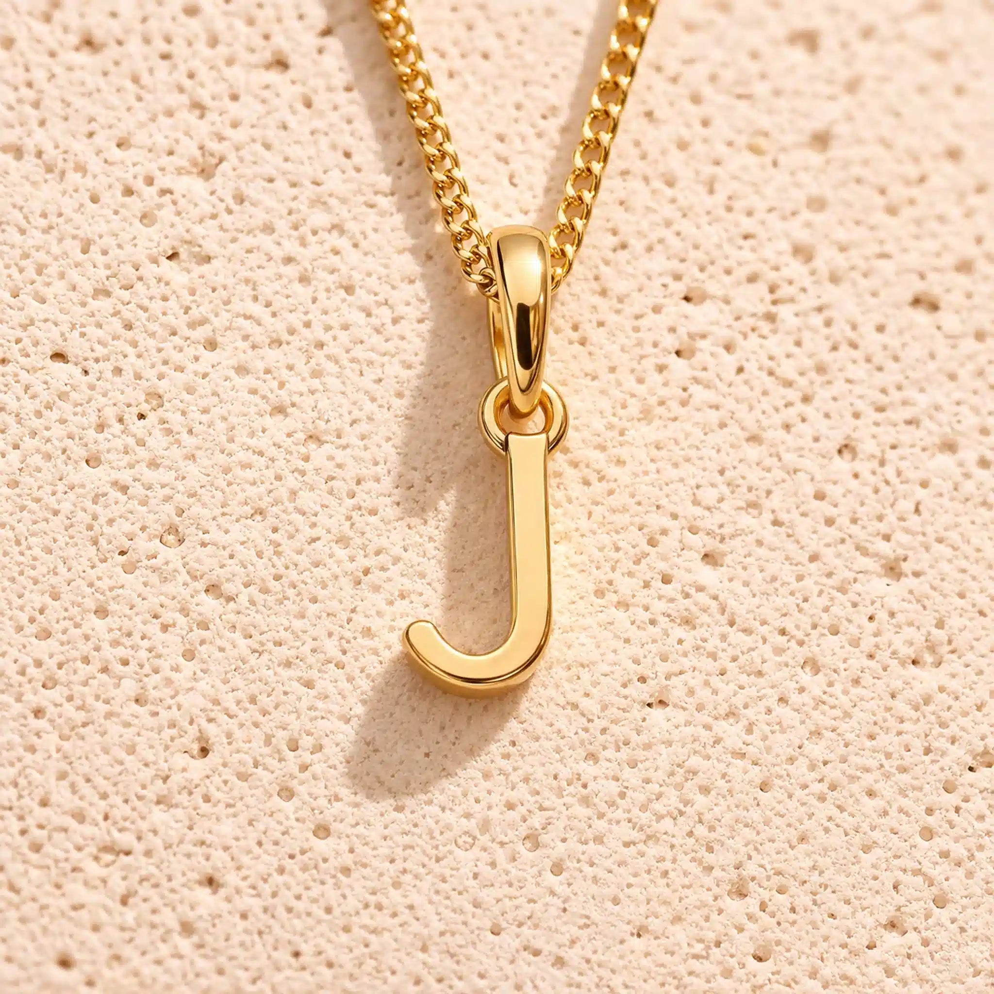 Personalised gold initial letter pendant necklace with polished uppercase J pendant on a fine cable chain in 9k gold plated sterling silver against a textured beige stone background