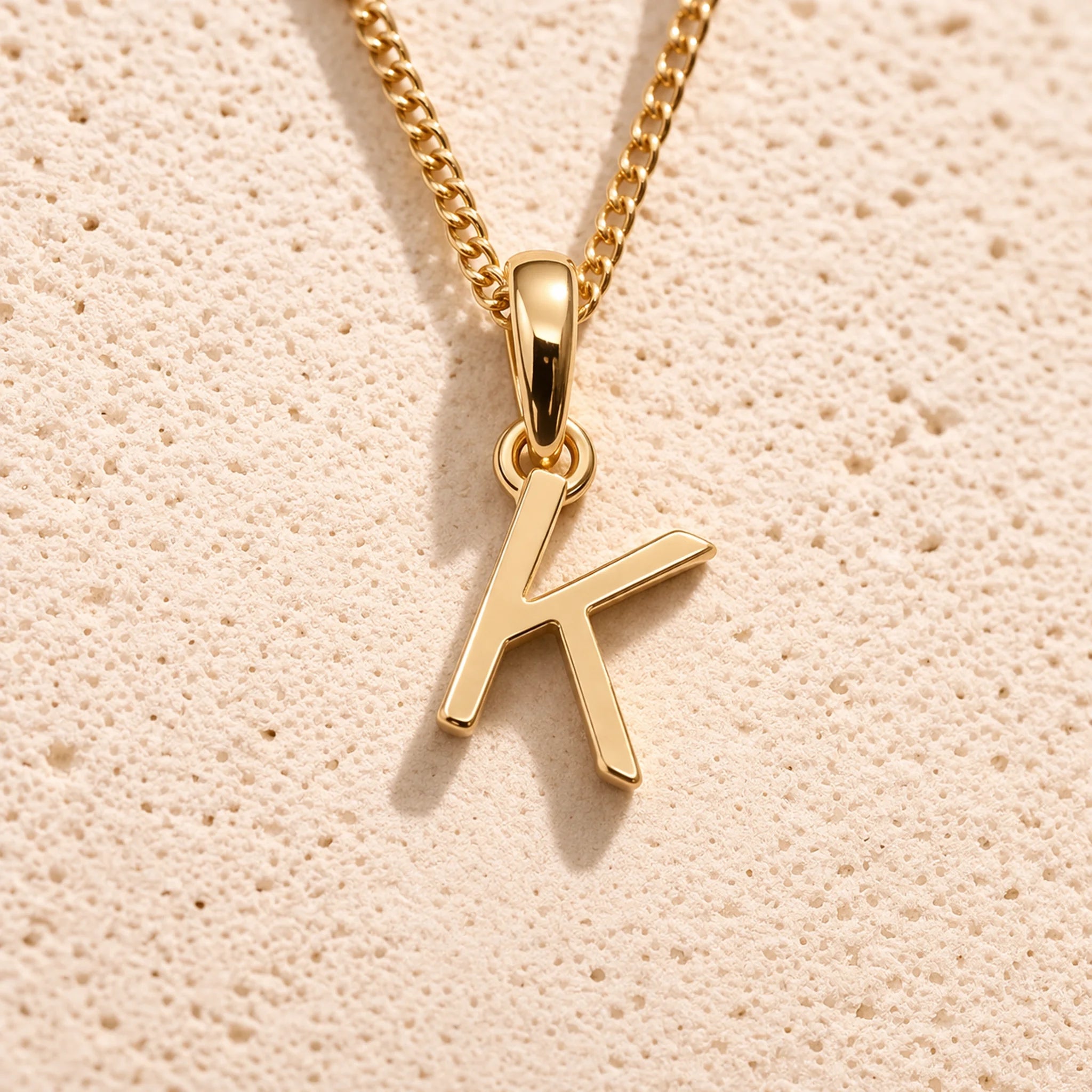 Personalised gold initial letter pendant necklace with polished uppercase K pendant on a fine cable chain in 9k gold plated sterling silver against a textured beige stone background