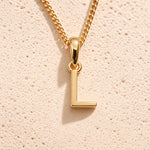 Personalised gold initial letter pendant necklace with polished uppercase L pendant on a fine cable chain in 9k gold plated sterling silver against a textured beige stone background