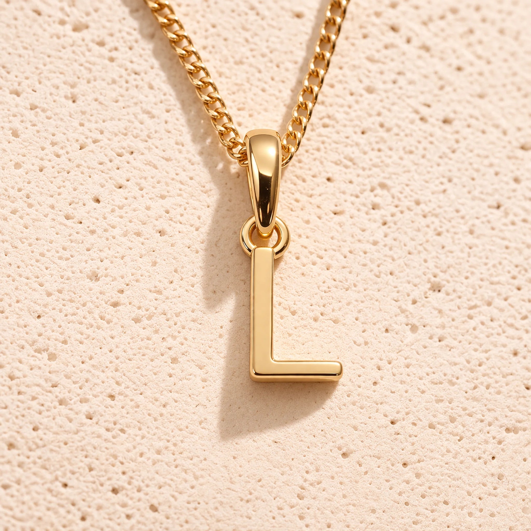 Personalised gold initial letter pendant necklace with polished uppercase L pendant on a fine cable chain in 9k gold plated sterling silver against a textured beige stone background