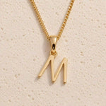 Personalised gold initial letter pendant necklace with polished uppercase M pendant on a fine cable chain in 9k gold plated sterling silver against a textured beige stone background