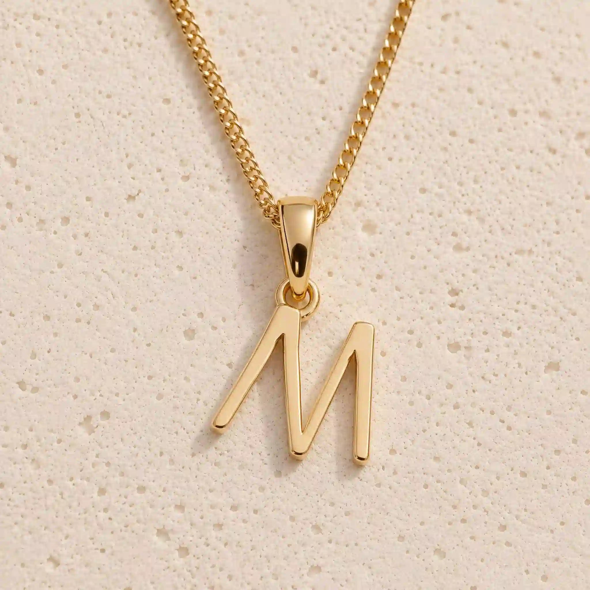 Personalised gold initial letter pendant necklace with polished uppercase M pendant on a fine cable chain in 9k gold plated sterling silver against a textured beige stone background