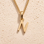 Personalised gold initial letter pendant necklace with polished uppercase N pendant on a fine cable chain in 9k gold plated sterling silver against a textured beige stone background