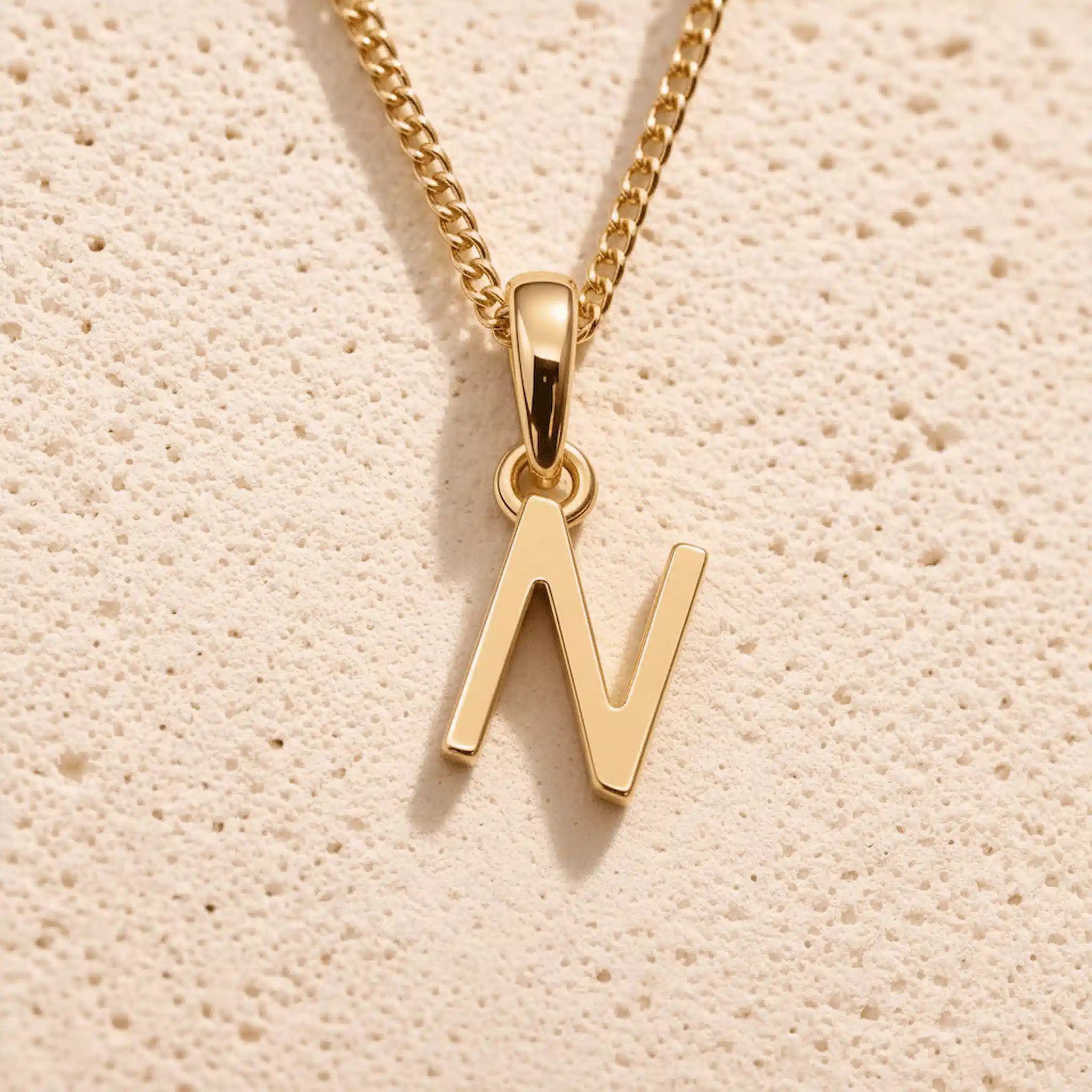 Personalised gold initial letter pendant necklace with polished uppercase N pendant on a fine cable chain in 9k gold plated sterling silver against a textured beige stone background
