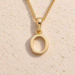 Personalised gold initial letter pendant necklace with polished uppercase O pendant on a fine cable chain in 9k gold plated sterling silver against a textured beige stone background