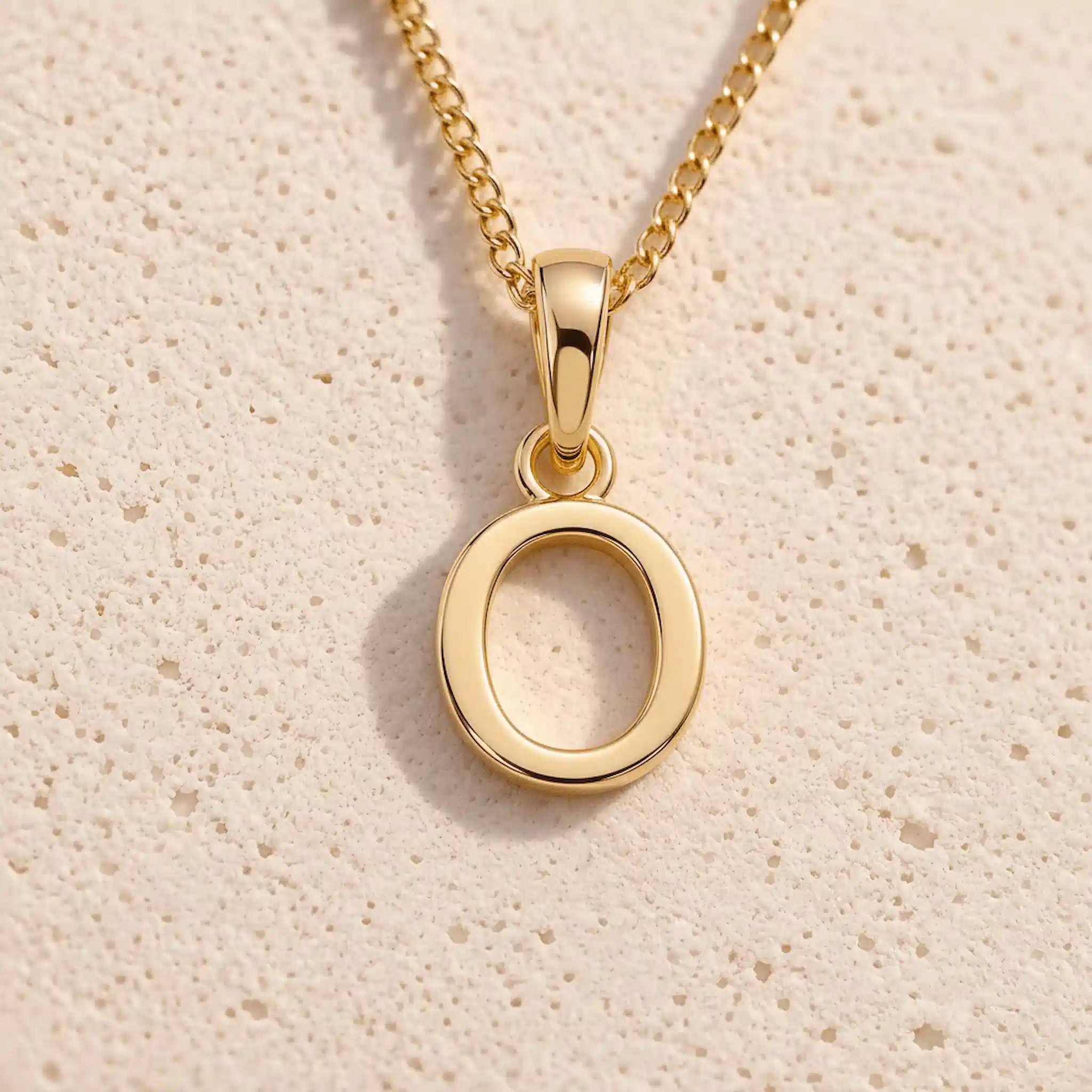 Personalised gold initial letter pendant necklace with polished uppercase O pendant on a fine cable chain in 9k gold plated sterling silver against a textured beige stone background