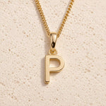 Personalised gold initial letter pendant necklace with polished uppercase P pendant on a fine cable chain in 9k gold plated sterling silver against a textured beige stone background