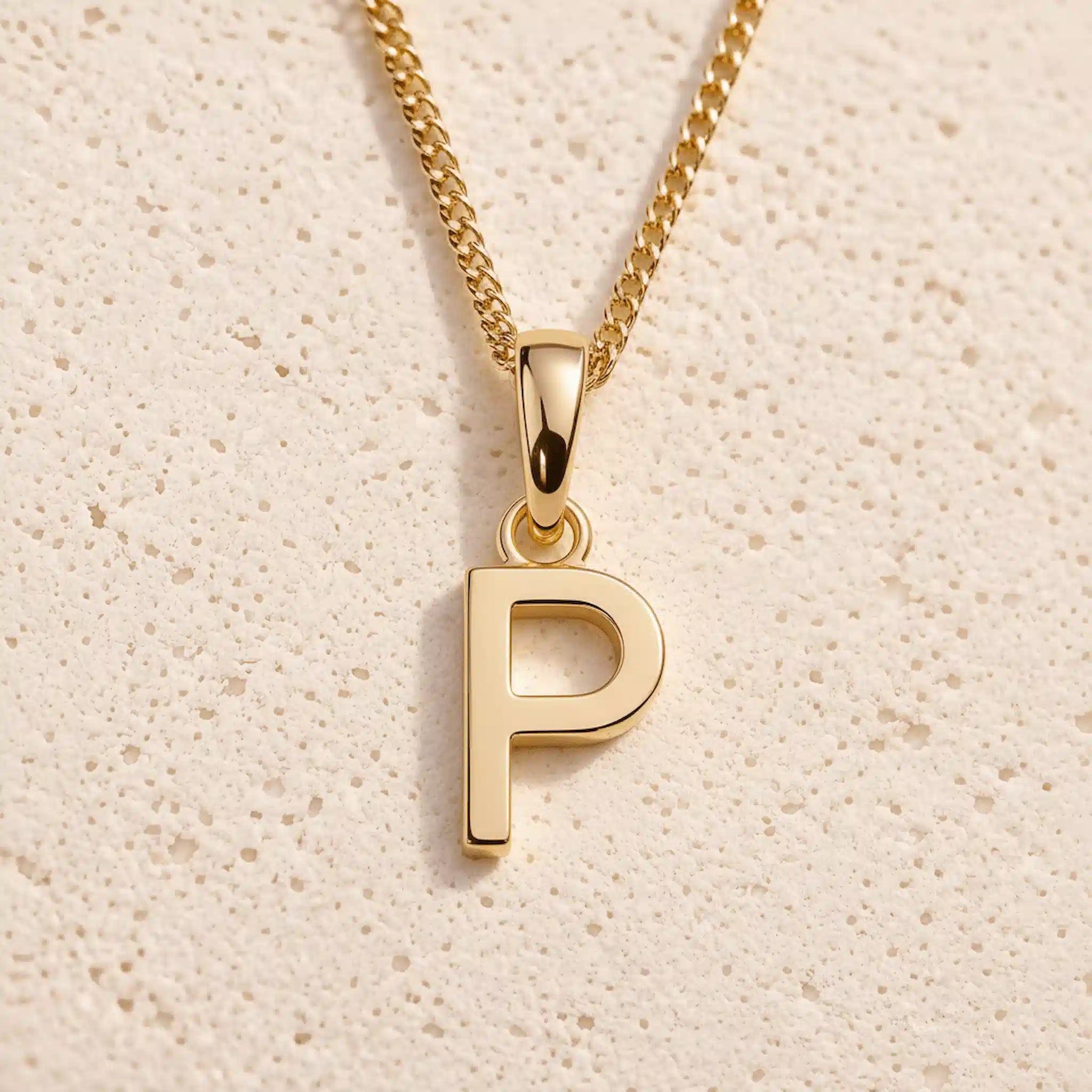 Personalised gold initial letter pendant necklace with polished uppercase P pendant on a fine cable chain in 9k gold plated sterling silver against a textured beige stone background