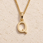 Personalised gold initial letter pendant necklace with polished uppercase Q pendant on a fine cable chain in 9k gold plated sterling silver against a textured beige stone background