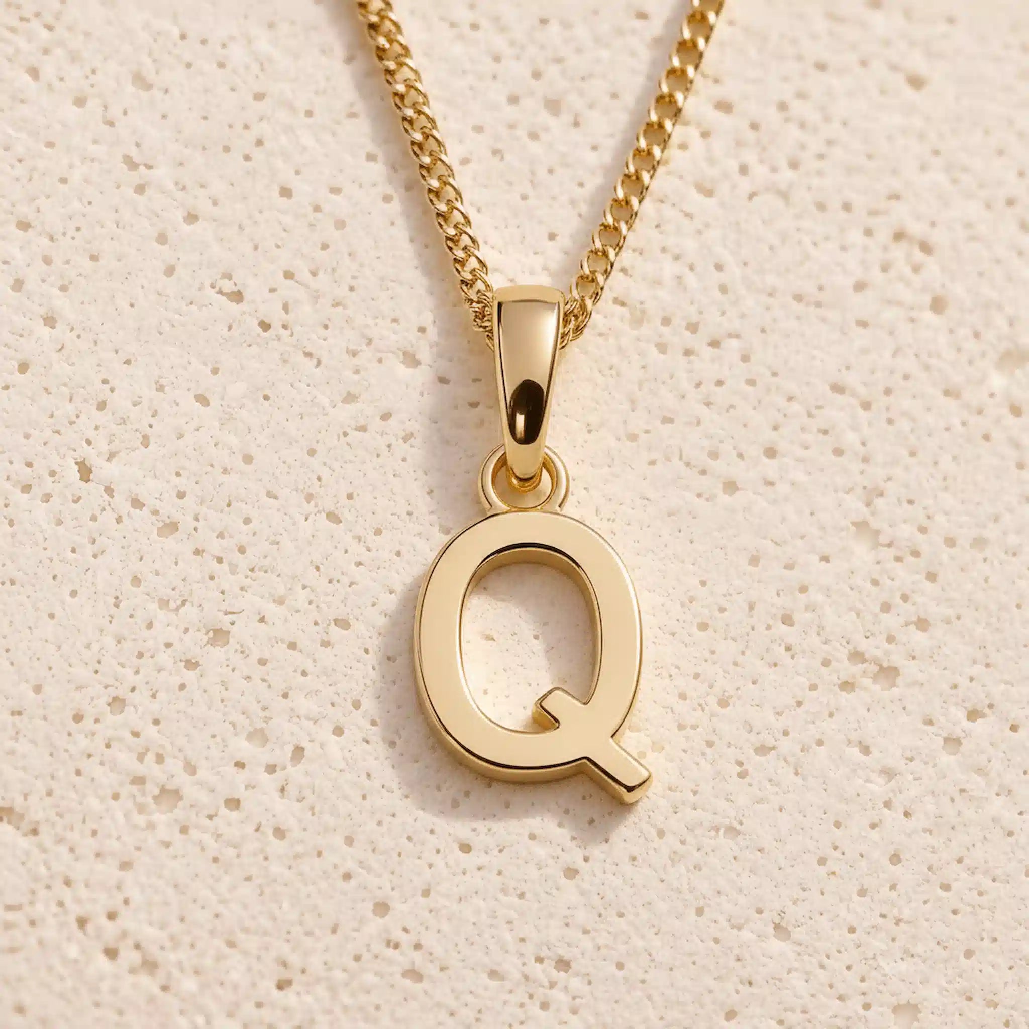 Personalised gold initial letter pendant necklace with polished uppercase Q pendant on a fine cable chain in 9k gold plated sterling silver against a textured beige stone background