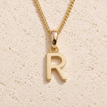 Personalised gold initial letter pendant necklace with polished uppercase R pendant on a fine cable chain in 9k gold plated sterling silver against a textured beige stone background