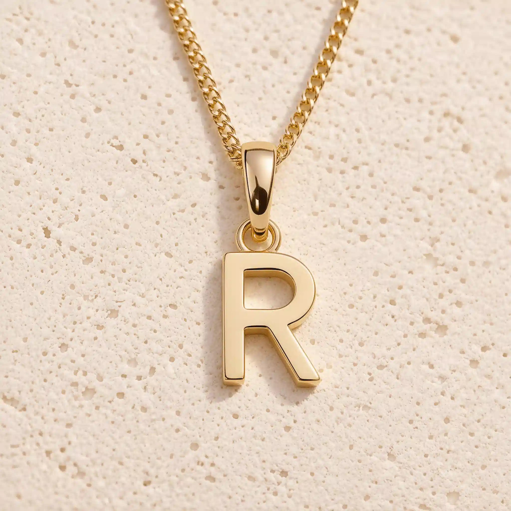 Personalised gold initial letter pendant necklace with polished uppercase R pendant on a fine cable chain in 9k gold plated sterling silver against a textured beige stone background