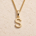 Polished gold initial pendant necklace with uppercase S charm on a fine link chain against beige stone