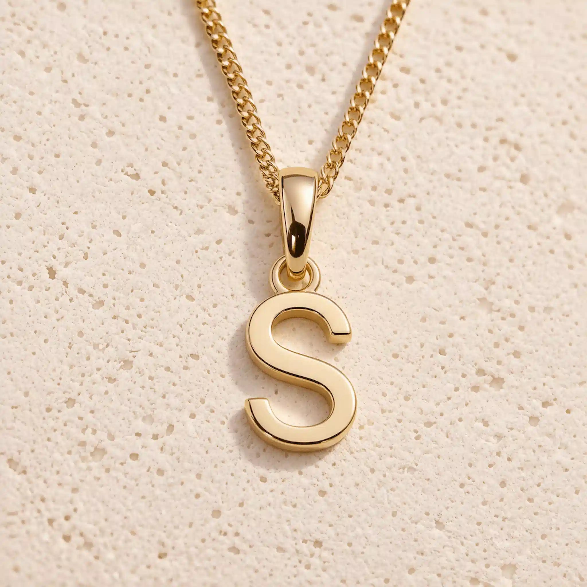 Polished gold initial pendant necklace with uppercase S charm on a fine link chain against beige stone