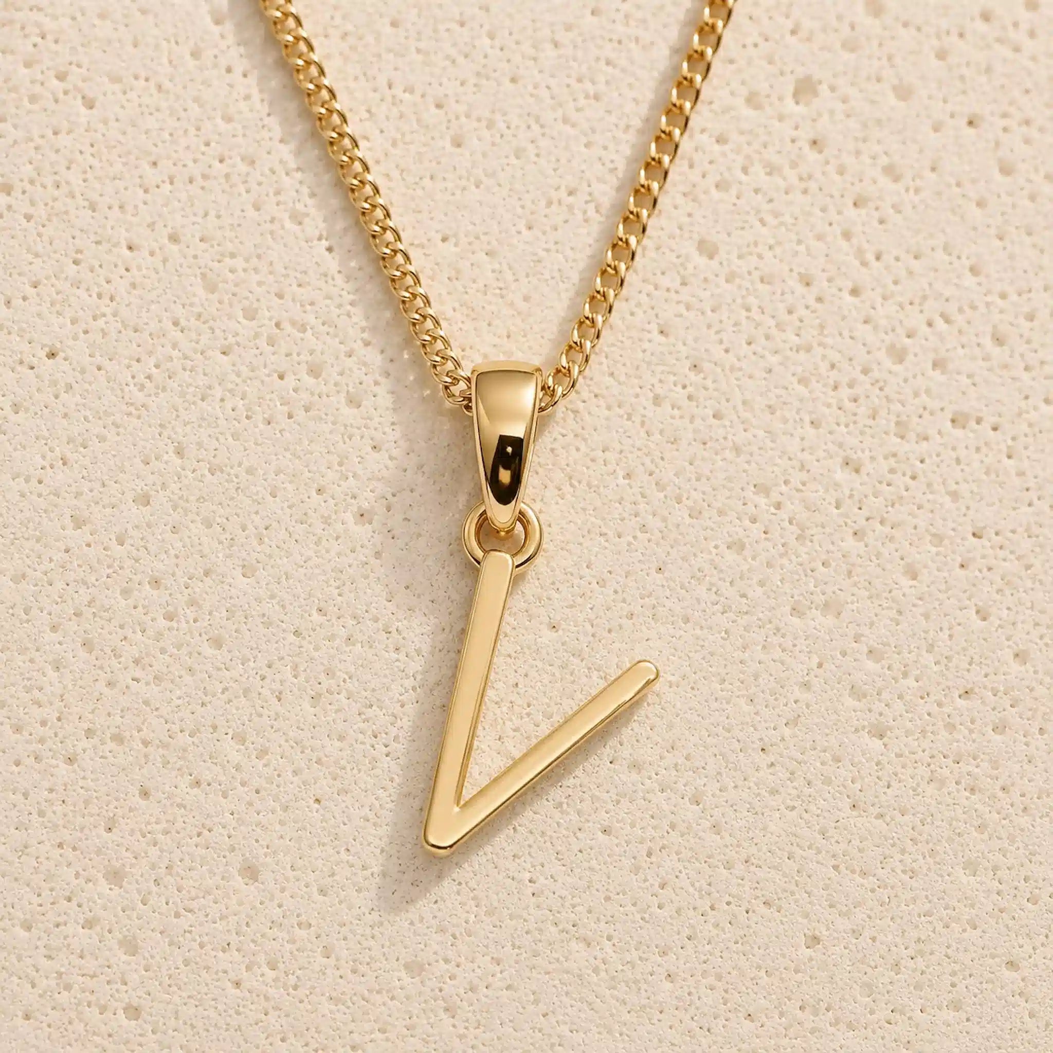 Polished gold initial pendant necklace with uppercase V charm on a fine link chain against beige stone