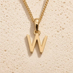 Polished gold initial pendant necklace with uppercase W charm on a fine link chain against beige stone