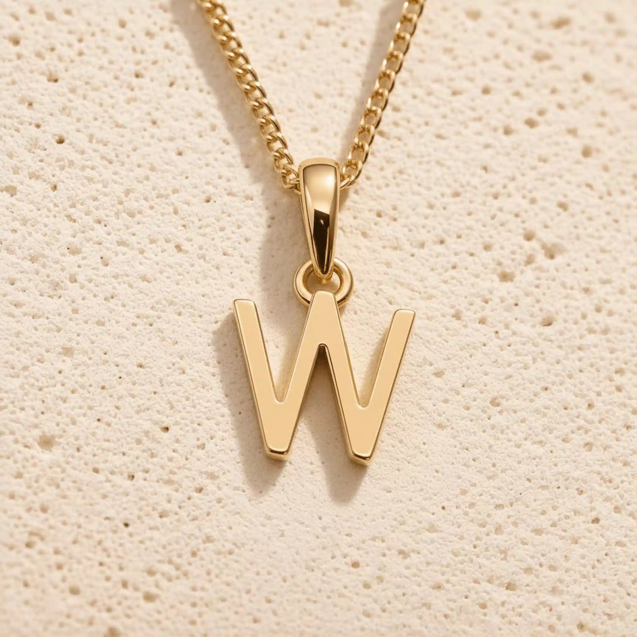 Polished gold initial pendant necklace with uppercase W charm on a fine link chain against beige stone