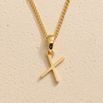 Polished gold initial pendant necklace with uppercase X charm on a fine link chain against beige stone
