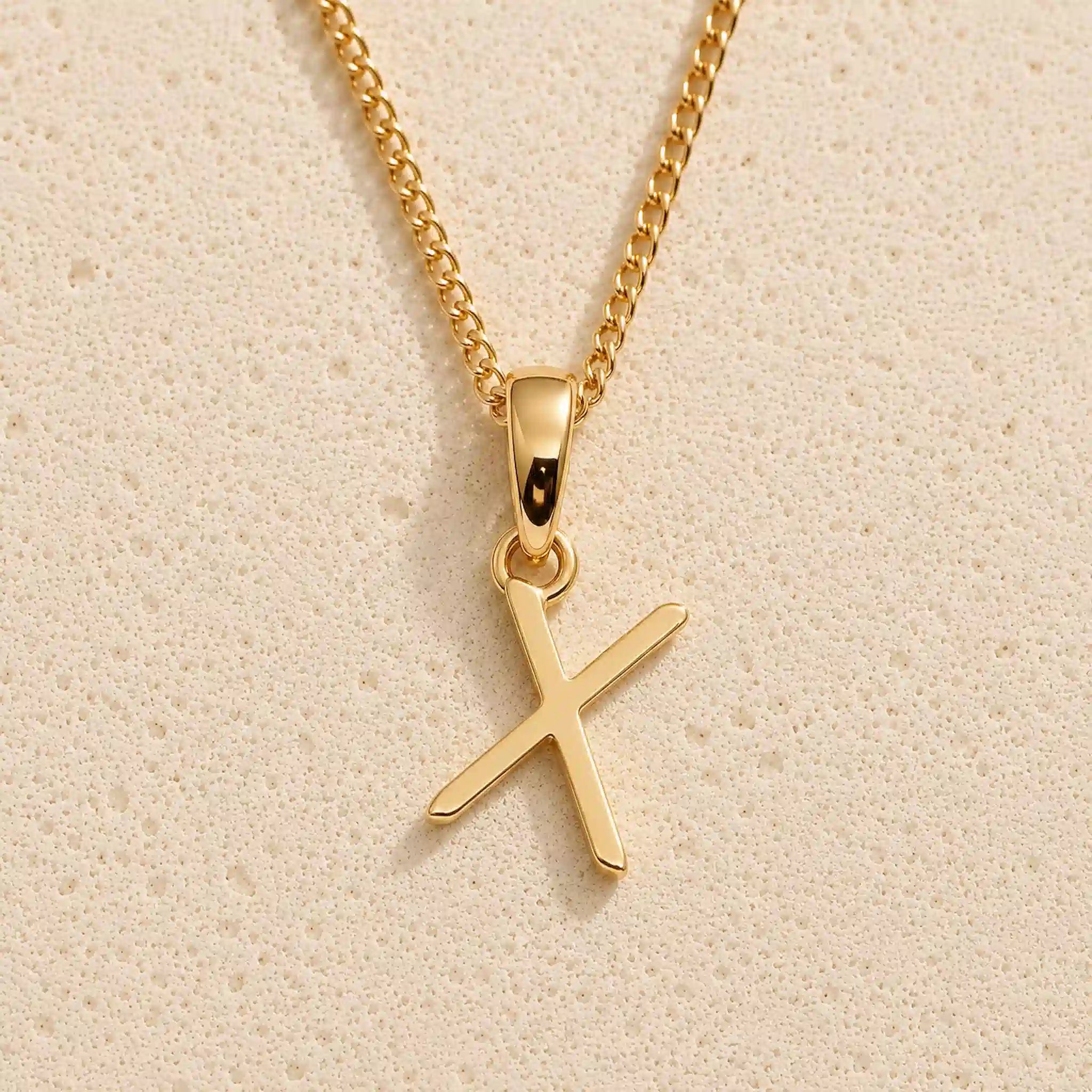 Polished gold initial pendant necklace with uppercase X charm on a fine link chain against beige stone