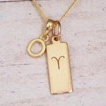 Gold initial O and Aries zodiac bar necklace, close-up of the letter charm and engraved pendant on whitewashed wood