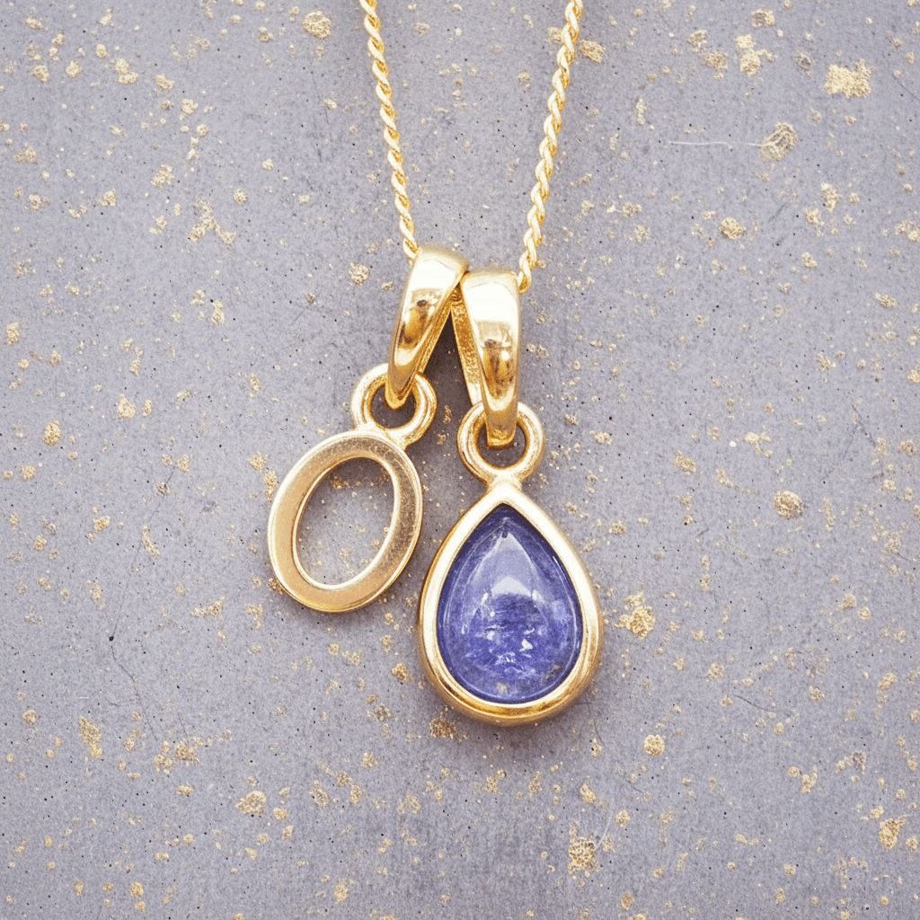 Gold necklace with a 'O' initial pendant and a purple teardrop december birthstone natural tanzanite gemstone pendant on a textured gray background