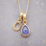 Gold necklace with a 'O' initial pendant and a purple teardrop december birthstone natural tanzanite gemstone pendant on a textured gray background