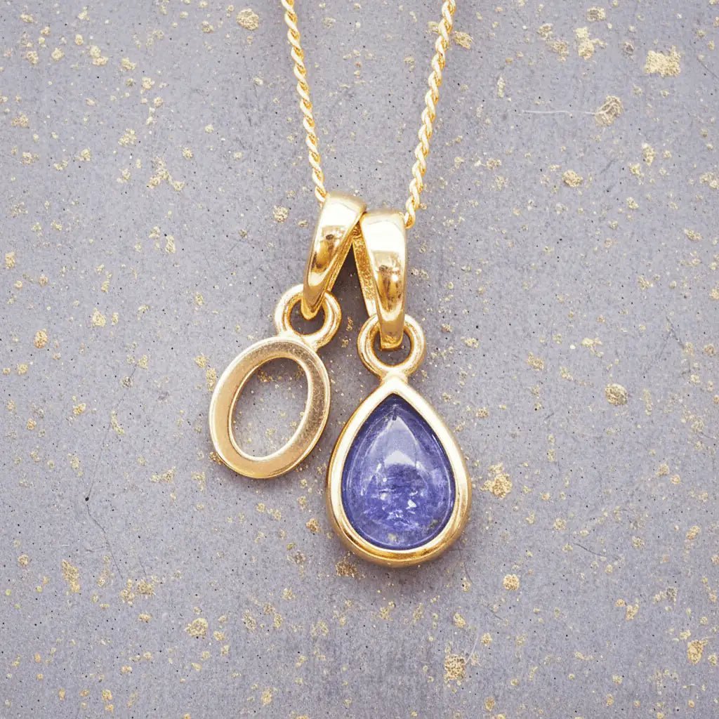 Gold December birthstone necklace with 'O' initial pendant and a natural tanzanite gemstone pendant on a grey background.