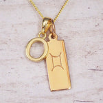 Gold initial O and Gemini zodiac bar necklace, close-up of the letter charm and engraved pendant on whitewashed wood