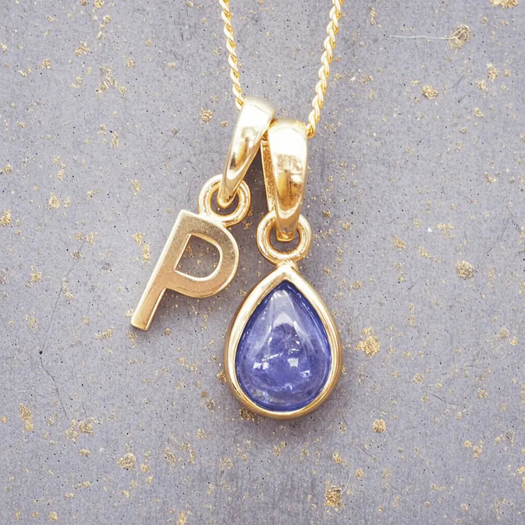 Gold December birthstone necklace with 'P' initial pendant and a natural tanzanite gemstone pendant on a grey background.