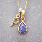 Gold December birthstone necklace with 'P' initial pendant and a natural tanzanite gemstone pendant on a grey background.