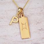 Gold initial P and Gemini zodiac bar necklace, close-up of the letter charm and engraved pendant on whitewashed wood