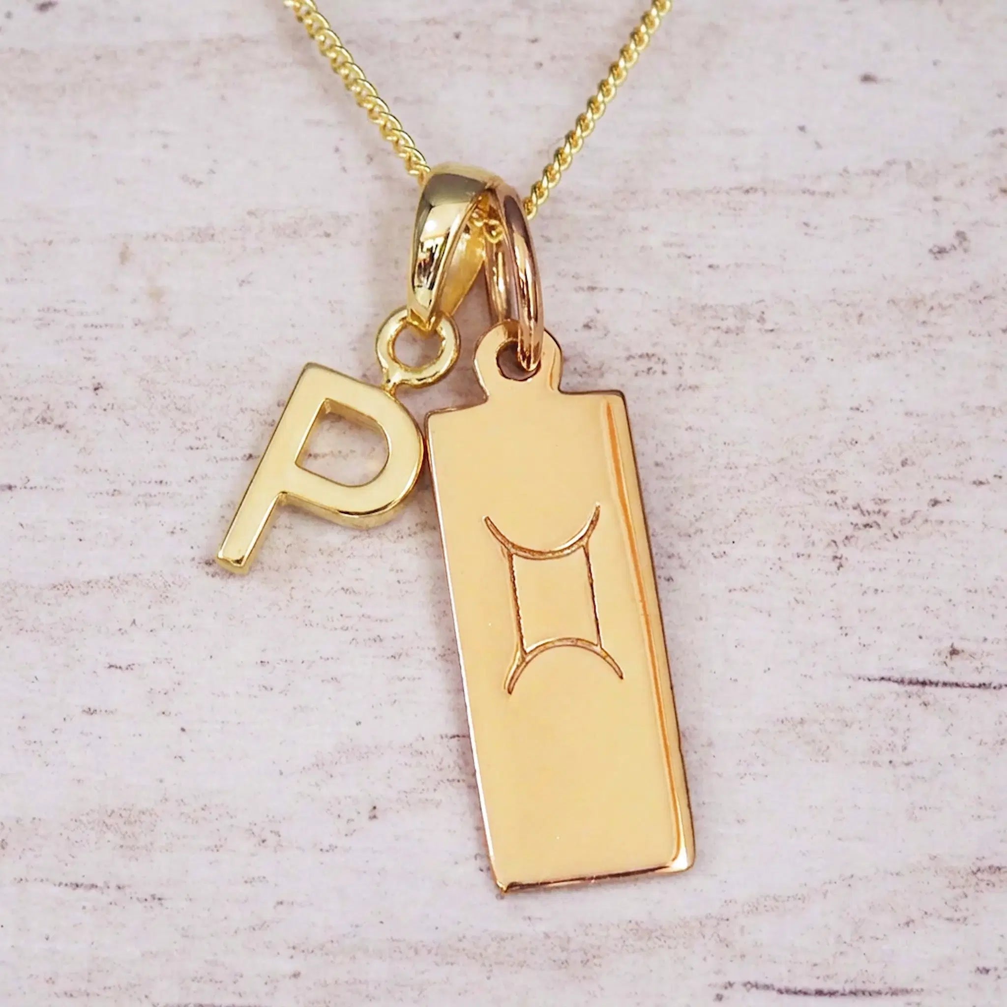 Gold initial P and Gemini zodiac bar necklace, close-up of the letter charm and engraved pendant on whitewashed wood